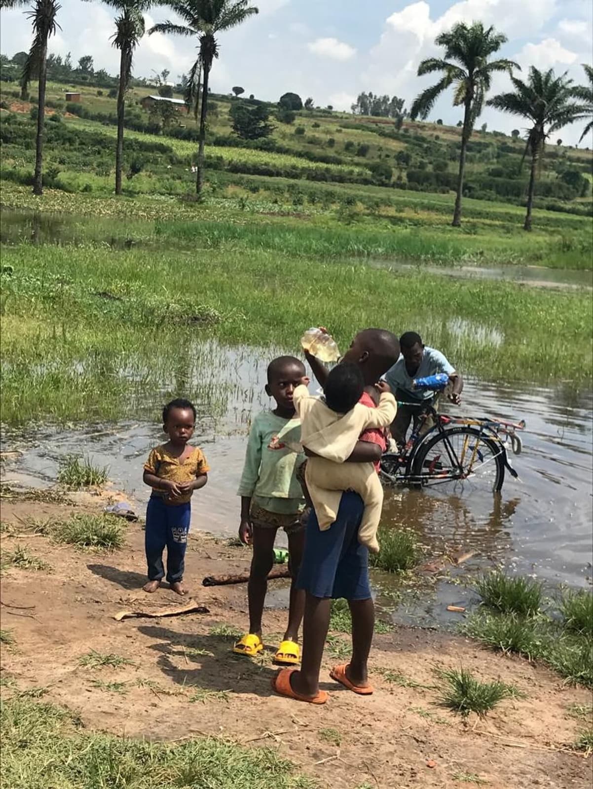 Children beside a shallow water source in Gashora, one holding a plastic bottle of untreated water, with a man washing a bicycle in the same water behind them.